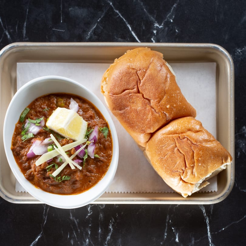 Tulsi Kitchen's Pav Bhaji, a vibrant Indian vegetable curry with soft bread rolls, lemon, and onions.