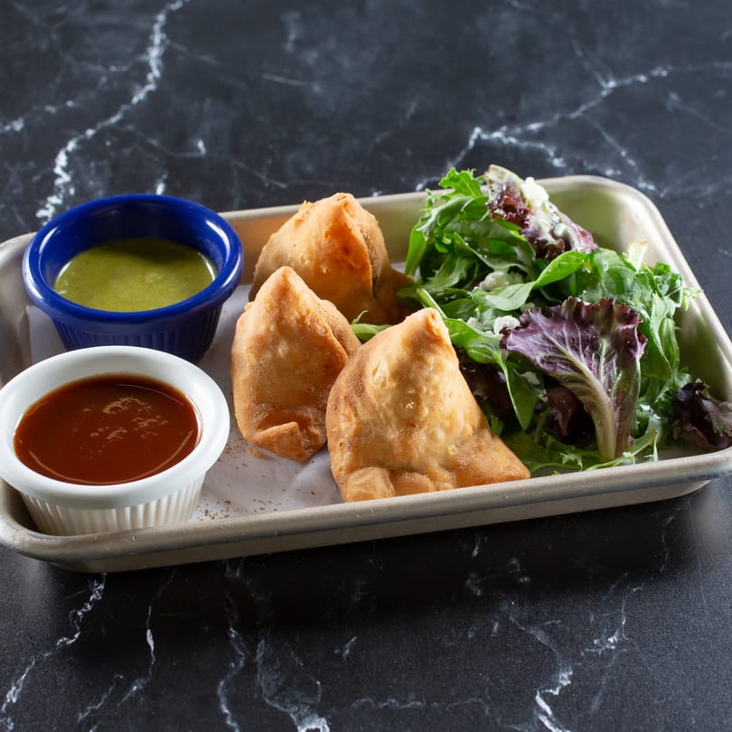 Three golden-brown samosas, two dipping sauces, and a fresh green salad on a tray.