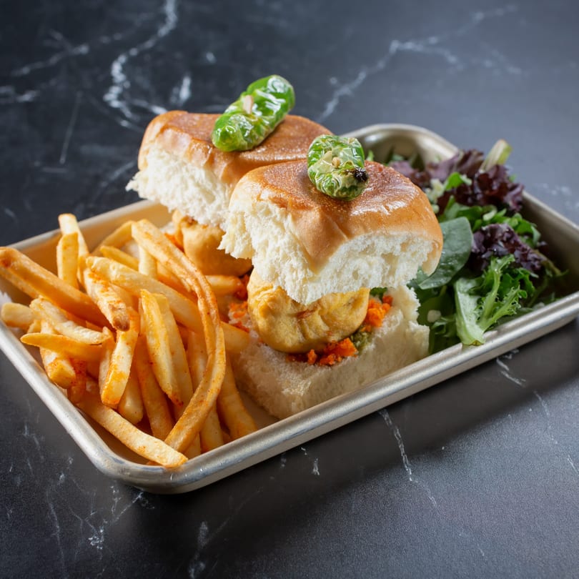 Two Indian vada pav sliders with green chili, fries, and salad on a metal tray.